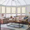 Pleated cellular blinds covering conservatory roof and bay windows, partially lowered and filtering soft daylight; in a bright, cozy sunroom with sofa, coffee table, woven rug, and minimal outside view.