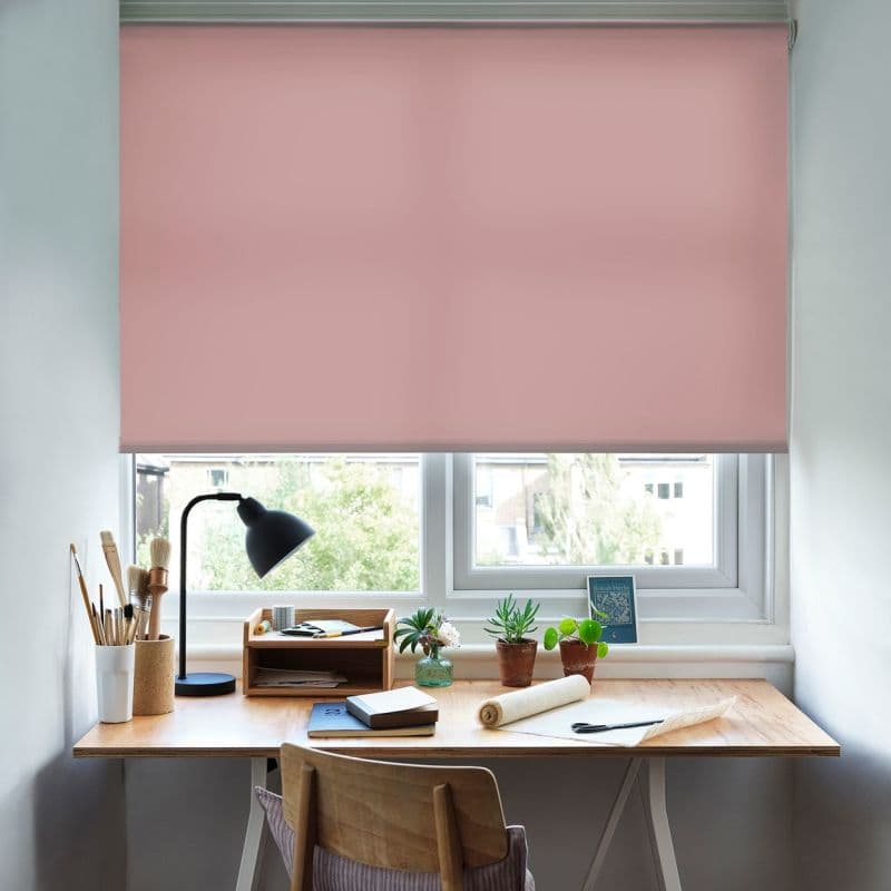 Pink roller blind, fully lowered with a slim lower gap, gently filtering daylight. Home workspace: wooden desk with black lamp, art brushes, notebooks and potted succulents beneath a double window.
