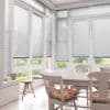 Horizontal aluminum Venetian blinds on tall bay windows, lowered to mid-height and filtering soft daylight, in a bright dining nook with a white table, chairs, cushions, tiled floor, and garden view.