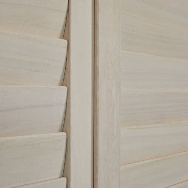 Pale wooden louvered shutters with a central vertical stile; left panel's slats are partially angled, right panel's slats closed, softly filtering interior light in a close-up of smooth wood grain.