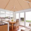 Beige pleated roof blinds and white pleated window/door blinds, fully lowered, filtering soft daylight; in a bright conservatory dining area with wooden table, upholstered chairs, sink and garden patio view.