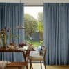 Blue pleated full-length curtains, hung and drawn slightly apart to reveal a glass patio door, framing a sunlit backyard; dining table with pendant lamp, pancakes and wooden chairs in foreground.