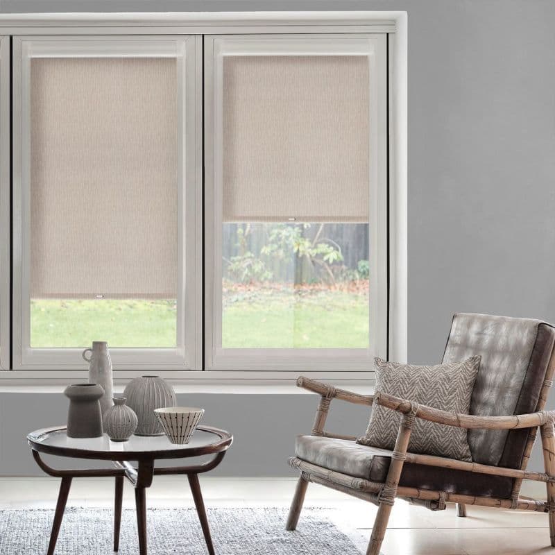 Beige fabric roller blinds (two adjacent panels, right slightly raised) covering a large window, filtering soft daylight into a grey-walled living room with rattan armchair and coffee table, garden view.