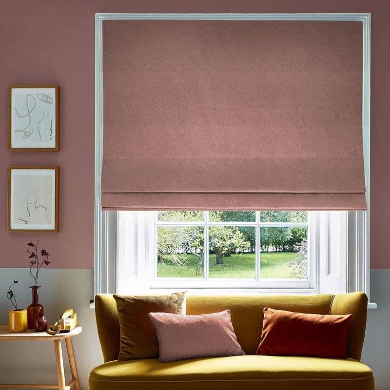A textured dusty-pink Roman blind is lowered over a white-framed sash window, partially filtering daylight in a living room with a mustard-yellow sofa, cushions, side table and green garden view.