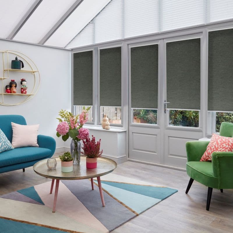 Dark gray roller blinds across four tall window/door panels, lowered to leave narrow bottom panes exposed, blocking light; set in a bright conservatory/sunroom with colorful seating, plants, and garden view.