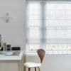 Semi-sheer, light-gray Roman shade with horizontal folds, lowered over a multi-pane sash window, diffusing pale daylight and obscuring leafless trees outside; minimalist home office with white desk, wooden chair.