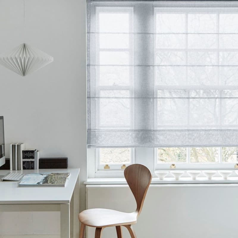 Semi-sheer, light-gray Roman shade with horizontal folds, lowered over a multi-pane sash window, diffusing pale daylight and obscuring leafless trees outside; minimalist home office with white desk, wooden chair.