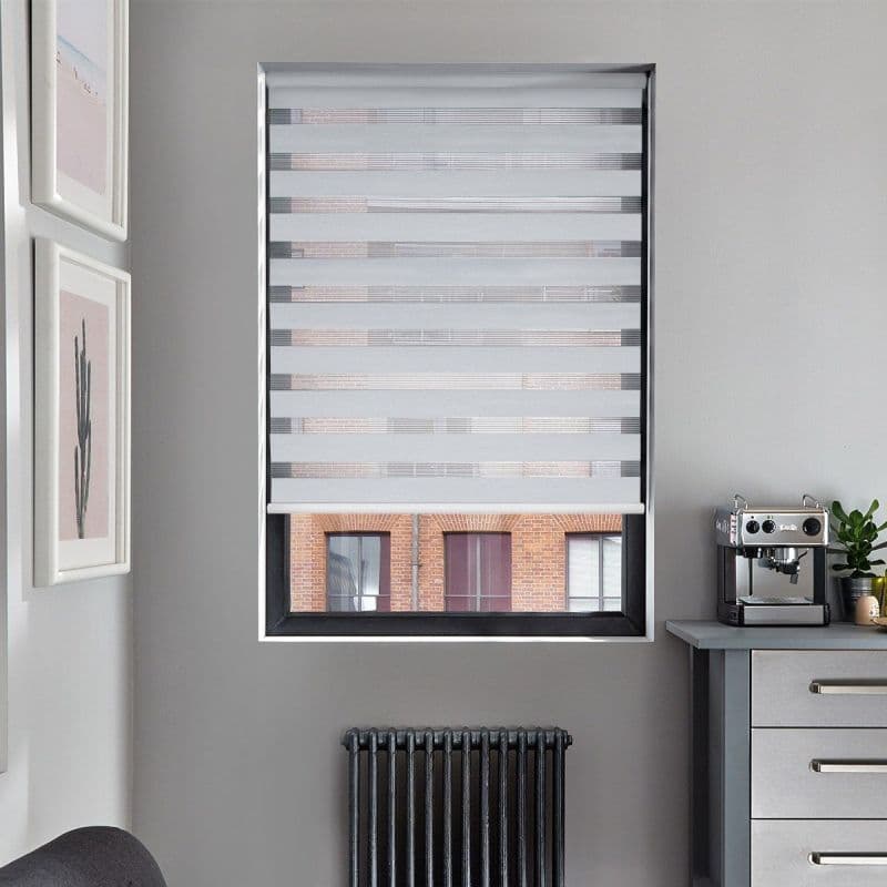 Zebra roller blind, lowered with alternating opaque-and-sheer horizontal stripes, filtering daylight and revealing a slit of exterior brickwork through the bottom; neutral kitchen corner with espresso machine, radiator, framed art.