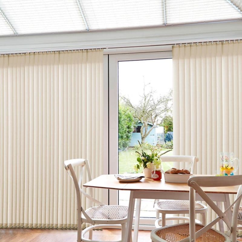 Beige vertical slatted blinds mounted at top, mostly closed but parted at center, filtering daylight; dining table and chairs with vase and pastries, garden shed and tree visible outside.