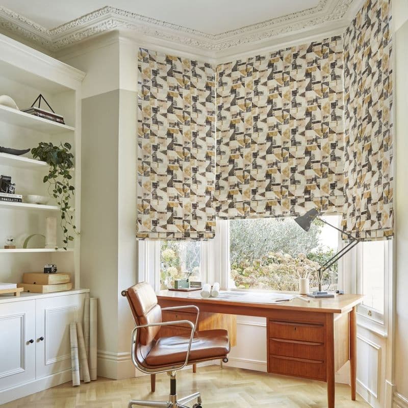 Patterned Roman blinds covering a bay window — geometric beige, gray and mustard print — partially lowered, filtering soft daylight; home office with mid-century wooden desk, leather swivel chair and built-in shelves, garden view.