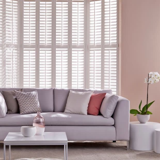 White louvered plantation shutters cover a tall bay window, closed and filtering daylight; in a pastel living room with light sofa, patterned and blush cushions, low coffee table and orchid.