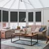 Dark grey cellular pleated blinds covering bay conservatory windows, lowered to mid‑height and blocking light; sunroom with beige sofa, chunky pink throw, wooden coffee table on blue rug.