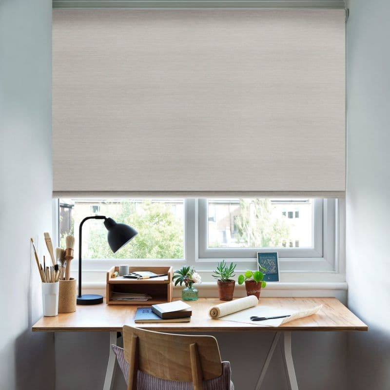Light beige roller blind lowered over a double window, diffusing daylight; below, a wooden desk with lamp, brushes, notebooks and small potted plants in a bright home office.