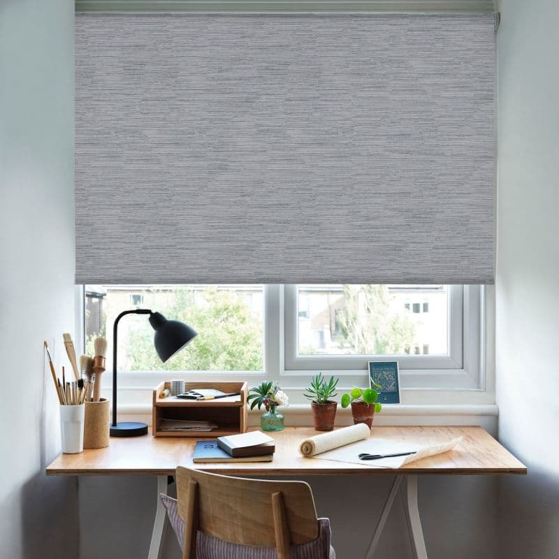 Grey textured roller blind lowered across a double window, largely blocking light; small home workspace below — wooden desk with lamp, paintbrushes, notebooks and potted succulents, soft daylight outside.