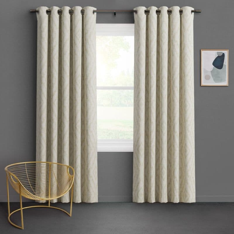 Grommet-top beige curtains, full-length panels on a rod, hung mostly closed with regular folds, filtering soft daylight over a white-framed window showing lawn and trees in a gray-walled room.