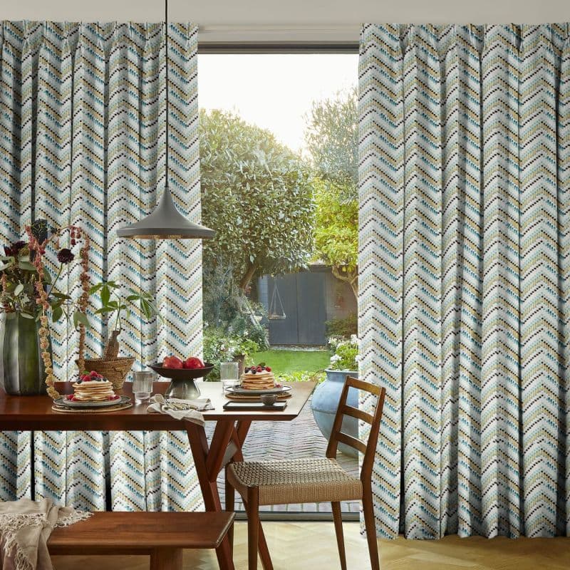Floor-to-ceiling chevron-patterned curtains hung at a glass door; partially drawn, framing the opening and admitting soft daylight; dining area with wooden table set and a sunlit garden beyond.