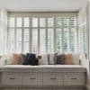 White plantation shutters cover tall bay windows, slatted and partially open to filter daylight; a built-in cushioned bench with patterned pillows and a tabby cat sits in a bright, airy room.