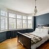 White plantation shutters on a bay window—five slatted panels with adjustable louvers partially open—filter soft daylight into a bedroom with dark blue walls, hardwood floor, black bed, and radiator.