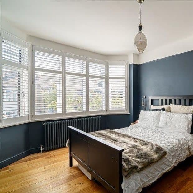 White plantation shutters on a bay window—five slatted panels with adjustable louvers partially open—filter soft daylight into a bedroom with dark blue walls, hardwood floor, black bed, and radiator.