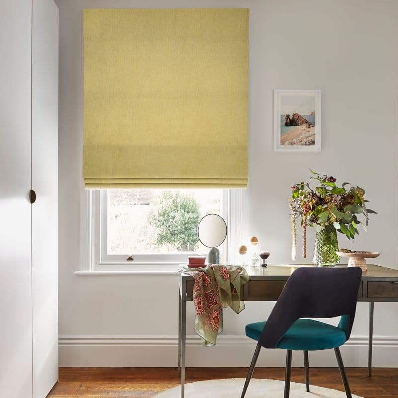 Yellow fabric Roman blind, lowered with soft folds, filtering daylight into a bright home office; desk beneath has a round mirror, vase of flowers, teal chair, and a small coastal print on the wall.