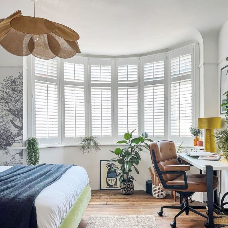 White plantation shutters on a five-panel bay window, closed and filtering daylight through angled slats, in a bright bedroom–home office with wooden floors, potted plants, bed, desk and leather chair.
