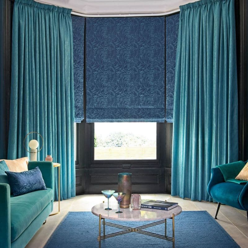 Teal floor-to-ceiling velvet curtains frame a lowered patterned navy Roman blind covering most of a bay window, filtering daylight. Cozy living room with teal sofas, marble coffee table, garden view.
IMPERFECT HOME