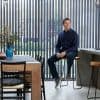 Floor-to-ceiling vertical blinds with narrow dark slats, mostly closed yet permitting garden views and daylight; modern kitchen/dining interior with wooden table, vase, man sitting on barstool smiling.