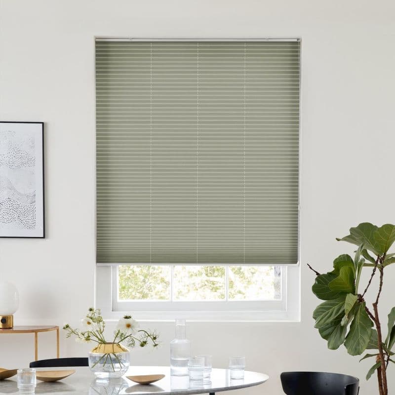 Sage-green pleated blind, fully lowered over a window with visible lower sash, filtering daylight; above a dining table with glassware, vase of daisies, framed art and a fiddle‑leaf fig.