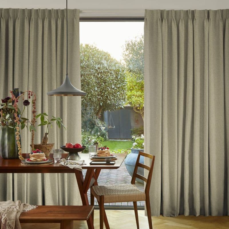 Full-length beige pleated curtains, drawn partly aside to reveal a central opening, framing and filtering soft daylight over a dining table set for breakfast and a green garden with trees and shed.