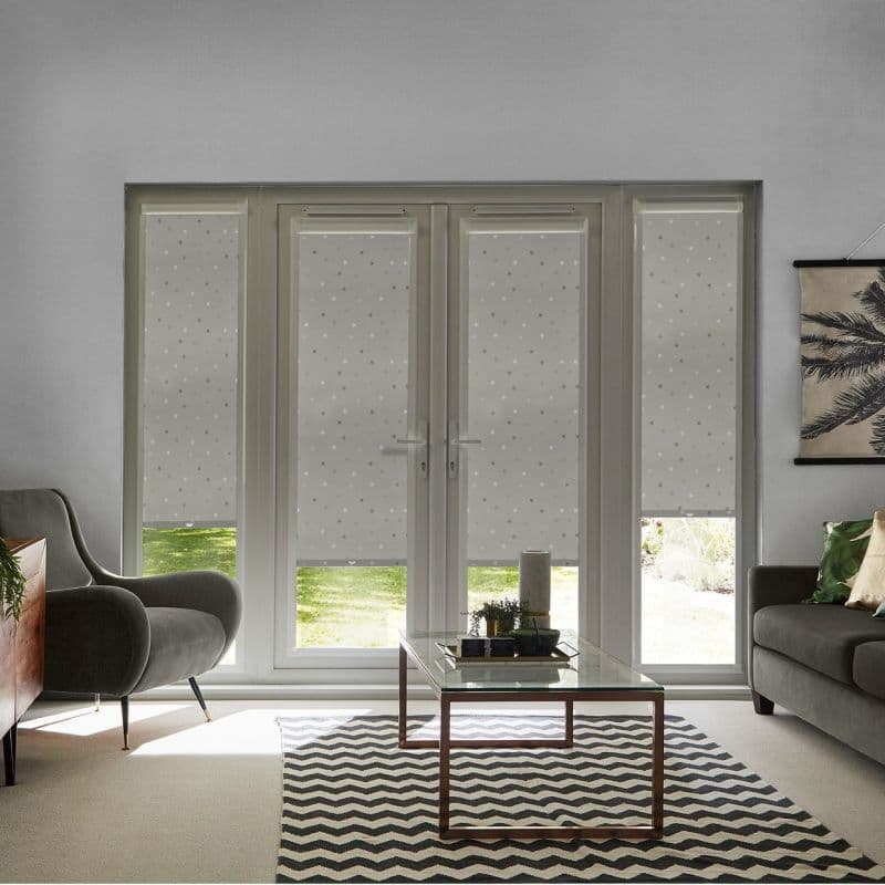 Star-patterned roller blinds covering French doors and sidelights, mostly lowered and filtering light, in a modern living room with armchairs, glass coffee table, chevron rug, and a sunlit garden.