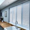 White plantation shutters covering a wide kitchen window, slats tilted to diffuse daylight; farmhouse sink and chrome faucet beneath; wooden countertop, muted grey walls and soft natural light.