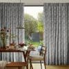Pleated grey patterned curtains frame a sliding glass door, partially drawn; they filter daylight into a dining area with a wooden table set for breakfast and a sunlit green backyard.