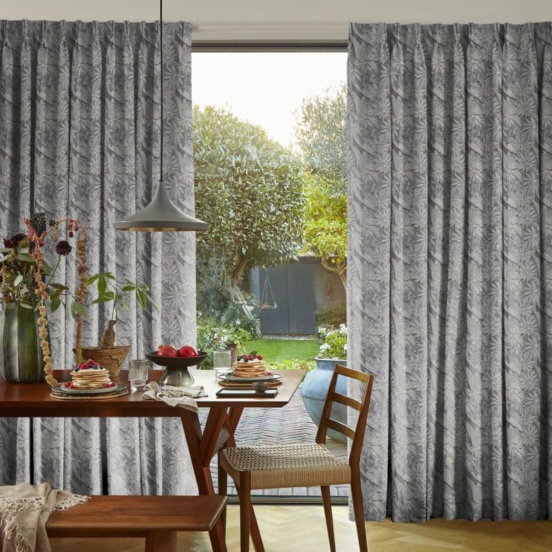 Pleated grey patterned curtains frame a sliding glass door, partially drawn; they filter daylight into a dining area with a wooden table set for breakfast and a sunlit green backyard.