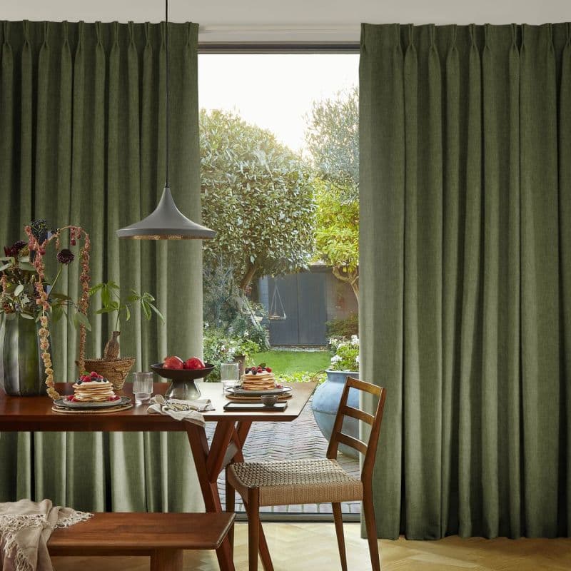Floor-length olive-green pleated curtains, drawn apart to expose the window, allowing soft daylight into a dining nook with a wooden table set for breakfast and a leafy garden view.