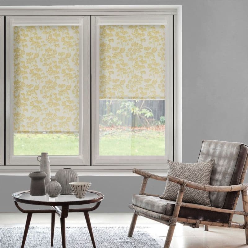 Floral-patterned roller blinds (yellow botanical print) covering two adjacent windows — left fully lowered, right partially lowered, filtering soft daylight; living room with gray walls, rattan leather chair and coffee table.