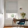 Grey textured Roman blind lowered across the window with soft folds, filtering daylight; a desk holding mirror, vase of flowers and teal chair in a neutral study overlooking a garden.