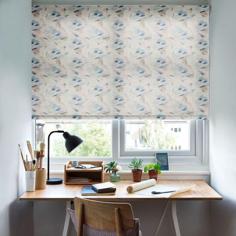Patterned roller blind, lowered across a double window—abstract pastel brushstroke print—filtering soft daylight; in front a tidy wooden desk with lamp, art brushes, small potted plants and a chair.