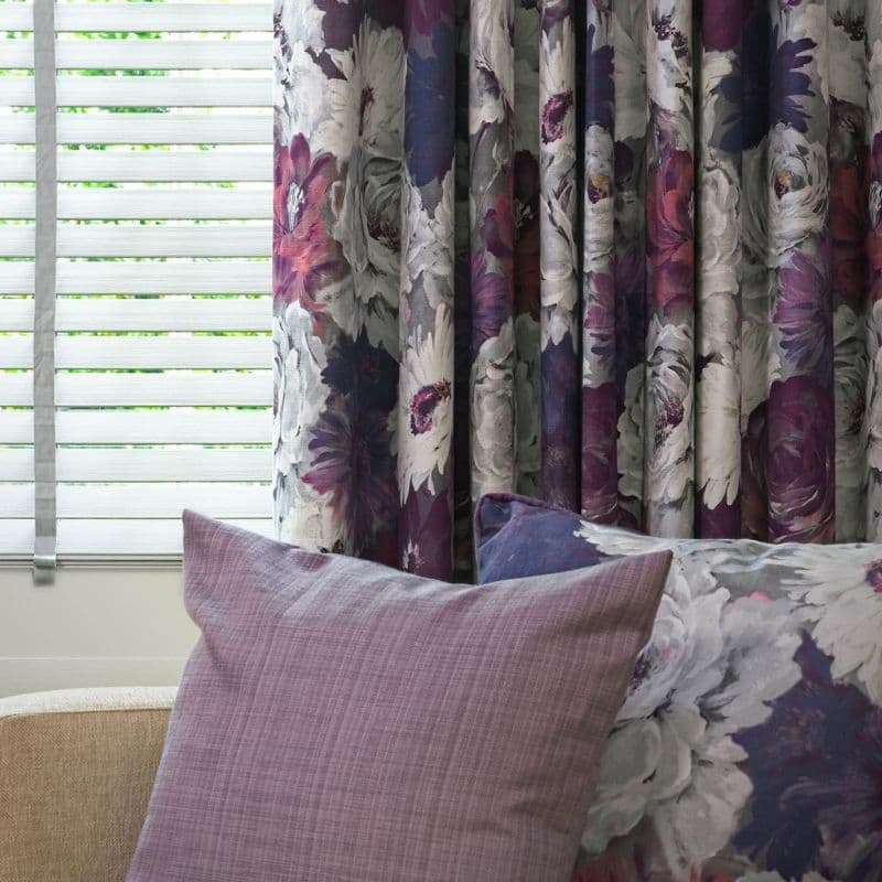 Floral curtains (purple, white peony pattern) hang pleated on the right, partially covering white horizontal blinds filtering daylight; mauve and floral cushions on nearby seating in a softly lit room.