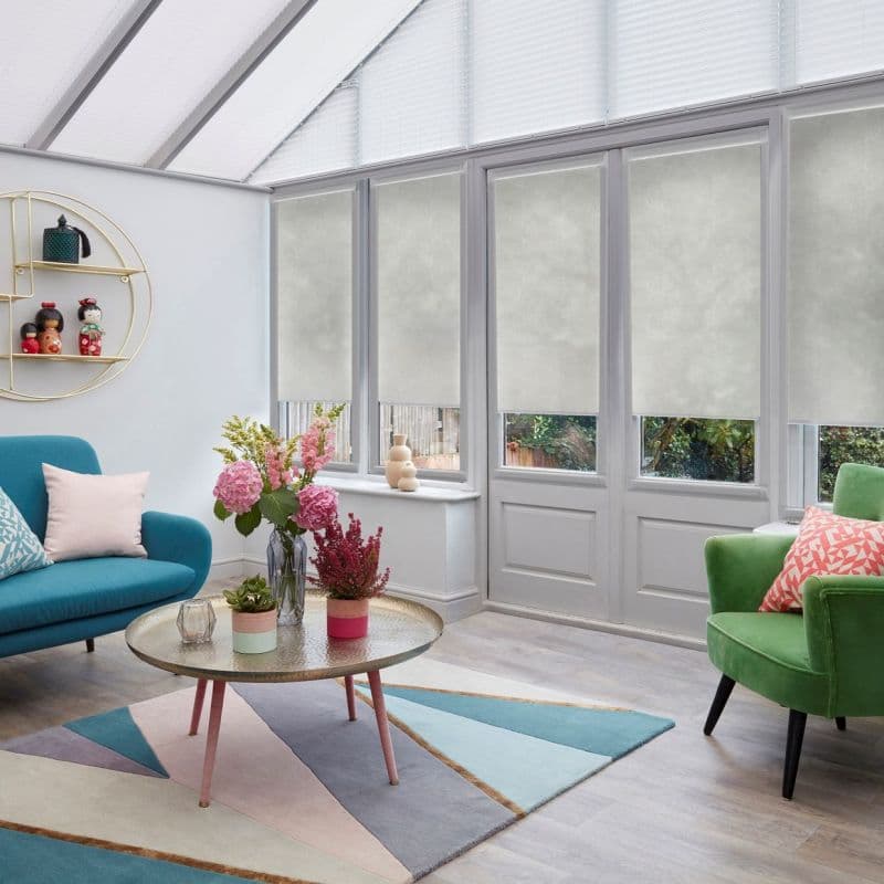 Light-gray roller blinds on conservatory windows and roof panes, mostly lowered to diffuse daylight and obscure the view; bright modern living room with colorful sofas, coffee table, and flowers.
