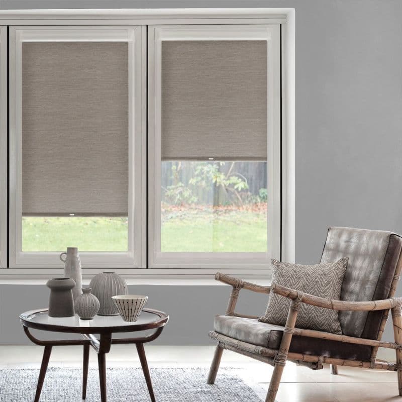Two beige fabric roller blinds, lowered to mid-height across a wide window, partially filtering daylight; living-room setting with gray walls, a rattan-framed leather armchair and a small coffee table.