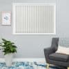 Light-gray vertical blinds, fully closed with beaded bottom chain, filtering soft daylight, in a calm living room with gray walls, potted plant, framed botanical print, gray armchair and blue rug.