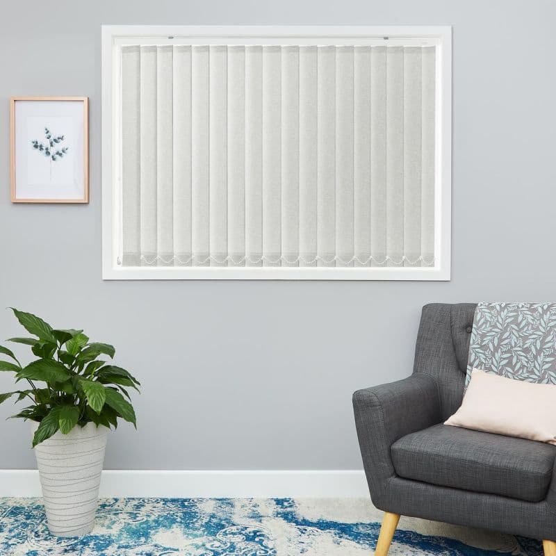 Light-gray vertical blinds, fully closed with beaded bottom chain, filtering soft daylight, in a calm living room with gray walls, potted plant, framed botanical print, gray armchair and blue rug.