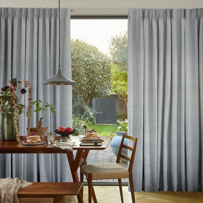 Pleated floor-length grey curtains, drawn open at the center, framing and filtering daylight through a glass door into a dining area with wooden table, pendant lamp, and sunlit garden beyond.