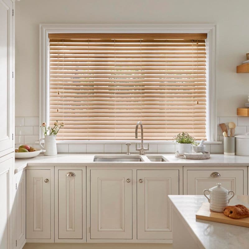 Horizontal wooden Venetian blinds covering a wide kitchen window; slats mostly closed, filtering soft daylight. In front, stainless sink, marble countertop, potted plant, crockery, pale cabinetry.