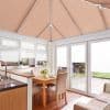 Peach pleated roof blinds and white pleated window and patio door blinds, all closed and softly filtering daylight in a kitchen-dining conservatory with table, chairs, and paved garden beyond.