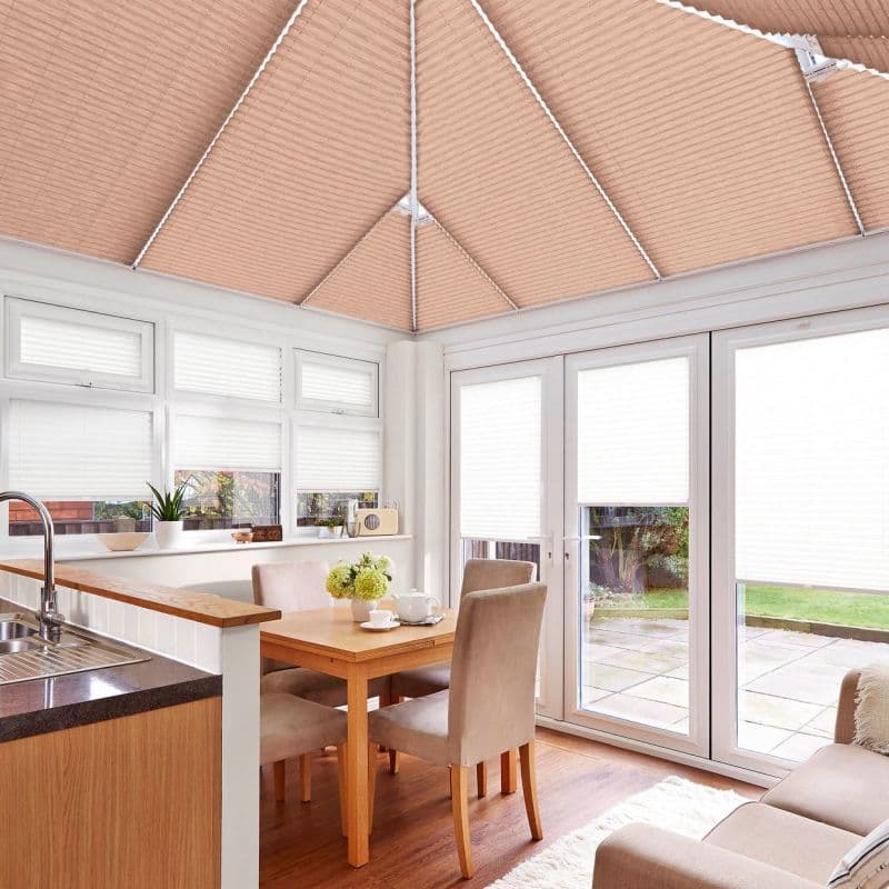 Peach pleated roof blinds and white pleated window and patio door blinds, all closed and softly filtering daylight in a kitchen-dining conservatory with table, chairs, and paved garden beyond.