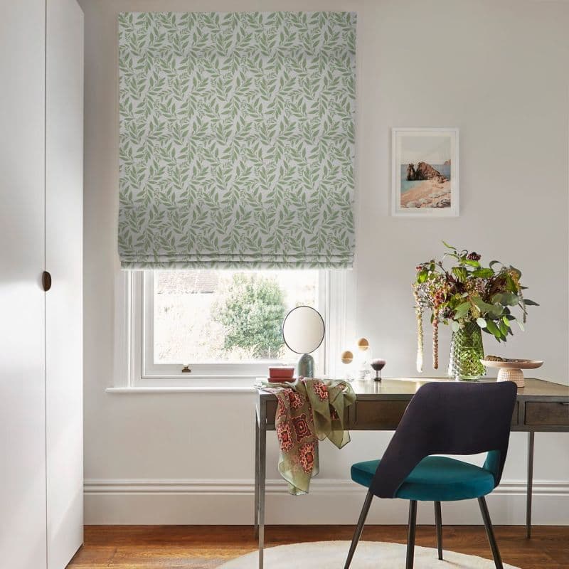 Leaf-patterned Roman blind lowered over a sash window, gently filtering daylight; framing a small home study with a desk, round mirror, vase of flowers, teal chair, and framed coastal print.
