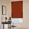 A rust-colored fabric roller blind, fully lowered and opaque, covering a tall window; filtering light into a minimalist home office with a black curved desk, wire chair, and pale walls.