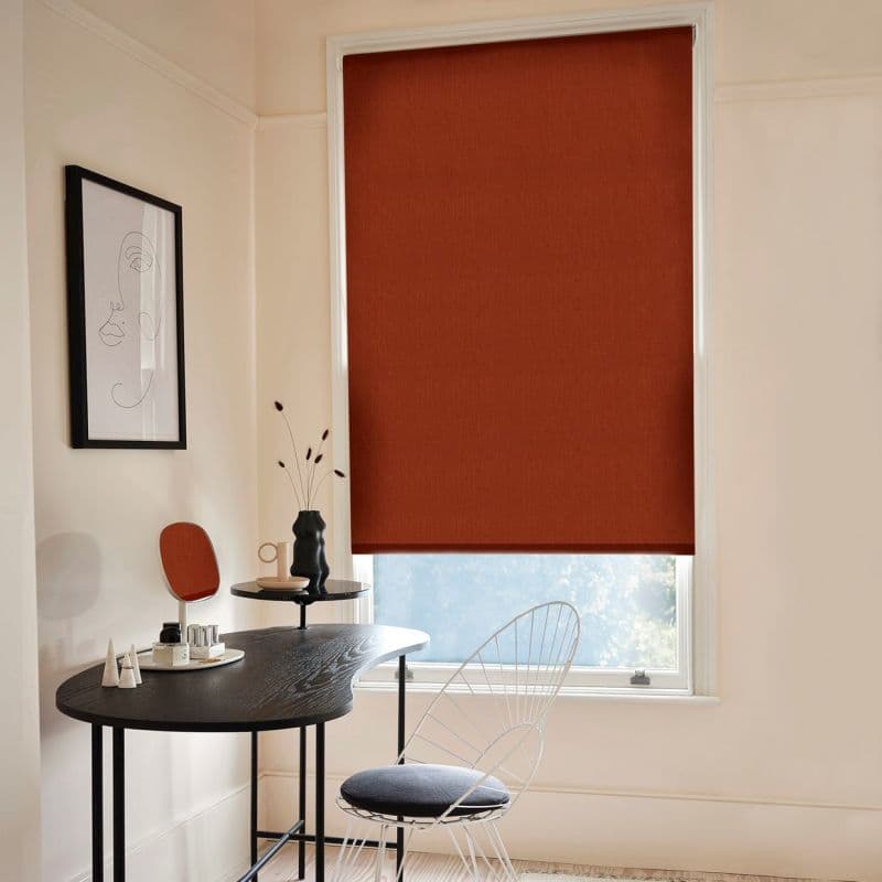 A rust-colored fabric roller blind, fully lowered and opaque, covering a tall window; filtering light into a minimalist home office with a black curved desk, wire chair, and pale walls.