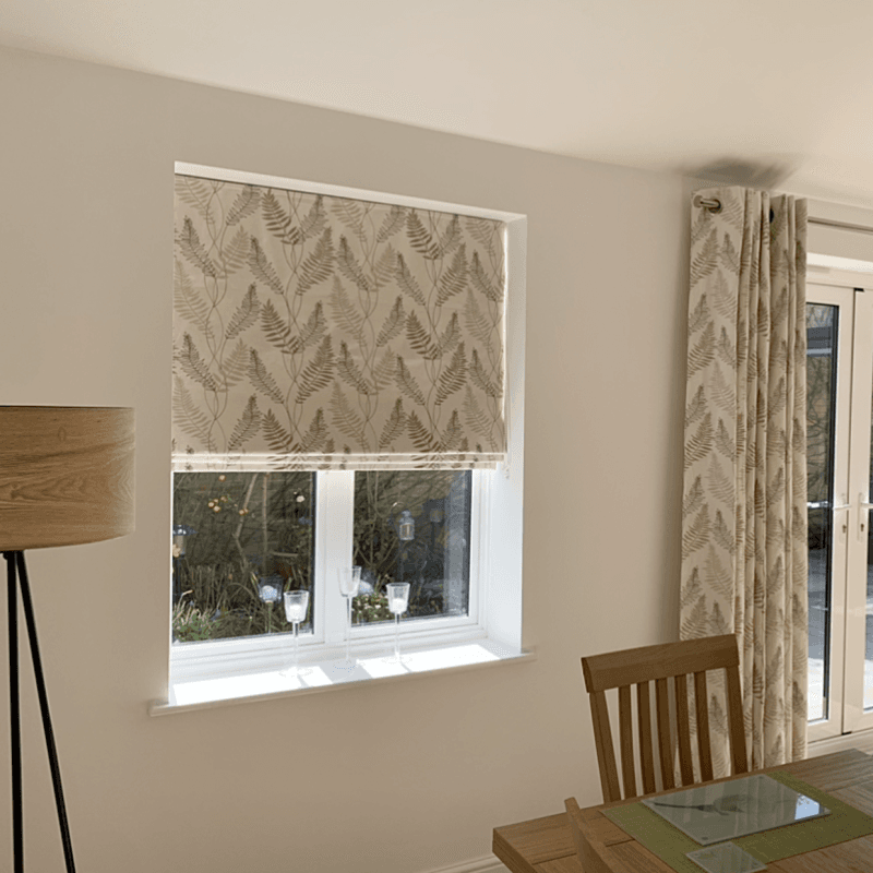 Patterned roller blind and matching grommet-top curtain, beige with fern print, lowered over a sash window (blind half-down) filtering daylight; dining area with wooden table, chair, lamp and garden view.